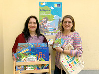 Projektmitarbeiterin Alexandra Marciniak und Projektleiterin Nicole Malina-Urbanz stehen vor einem Plakt von der Aktion "Lesepass 2022" und halten Bücher in den Händen