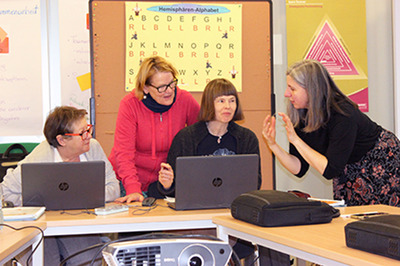 Eine Gruppe Teilnehmerinnen der Basisbildung sitzen vor zwei Laptops. Sie schauen gespannt zu einer Lernbegleiterin die ihnen gerade etwas erzählt.