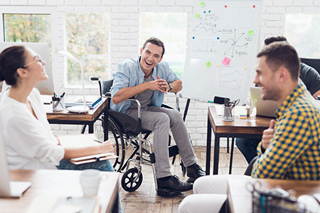 Eine Frau und zwei Männer, davon sitzt einer im Rollstuhl, diskutieren und lachen in einem modernen Büro.