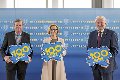 Landesrat Gottfried Waldhäusl, Landeshauptfrau Johanna Mikl-Leitner und LH-Stellvertreter Franz Schnabl stehen vor einer blauen Wand und halten das Logo von "100 Jahre Niederösterreich" hoch