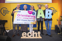 Gruppenfoto mit 5 Personen. Sie halten ein großes Schild in die Höhe mit einer Ankündigung zur BhW Wissens.Tour 2025