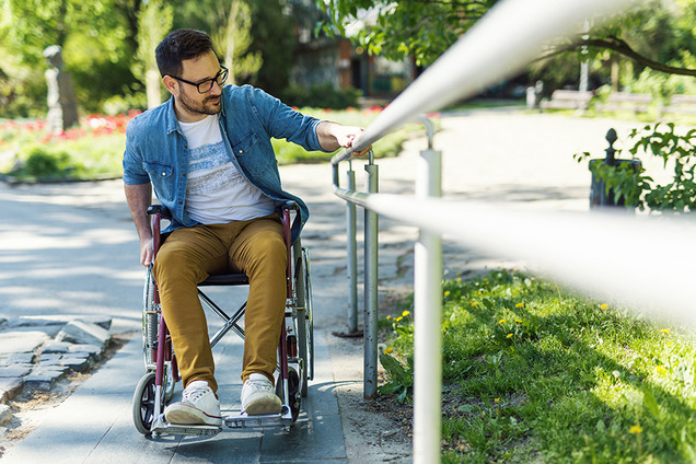 Ein Mann sitzt im Rollstuhl und versucht eine Rampe hochzukommen, indem er sich an einem Geländer fest hält.