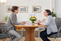 Zwei Frauen sitzen sich an einem runden Tisch in einem hellen Büro gegenüber und führen ein Beratungsgespräch. Im Hintergrund sind Bilder an der Wand, ein Blumenstrauß auf dem Tisch und Arbeitsmaterialien zu sehen, was eine ruhige, professionelle Atmosphäre vermittelt.