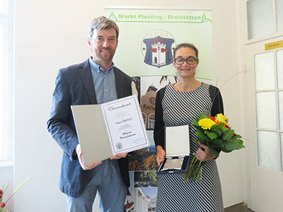 Herr Bürgermeister Roland Braimeier überreicht Bildungswerkleiterin Tatjana Nikitsch eine Urkunde und eine Medialle. Frau Nikitsch hält die silberne Ehrenplakette und einen Strauß Blumen in den Händen.