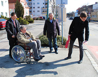 Drei Männer stehen, ein Mann sitzt im Rollstuhl, vor einem Zebrastreifen. Sie begutachten die Höhe der Randsteinkante, im Rahmen einer Gemeindebegehung. 