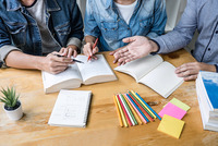 Auf einem Schreibtisch liegen aufgeschlagene Bücher und Stifte. Man sieht noch die Oberkörper und Amre von drei Personen die sich anscheinend angeregt unterhalten.