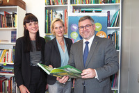 Gruppenbild, die drei Personen stehen vor vollen Bücherregalen und schauen gemeinsam in ein Buch
