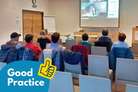 Mehrere Personen sitzen in einem Seminarraum und verfolgen eine Online-Präsentation, die auf eine Leinwand projiziert wird. Auf dem Bildschirm ist eine Frau im Videocall mit dem Logo von „BhW Niederösterreich“ zu sehen.