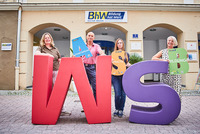 Vier Lernbegleiterinnen und Lernbegleiter der Basisbildung stehen vor dem Haus Linzerstraße 7. Davor stehen noch zwei sehr große Buchstaben aus Schaumstoff. Ein großes "W" in rot und ein großes "S" in lila. Drei der Personen halten auch kleinere Buchstaben in den Händen.