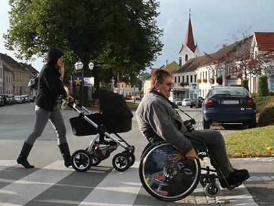 eine Frau mit Kinderwagen und ein Rollstuhlfahrer überqueren eine Straße auf einen Zebrastreifen