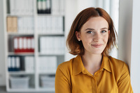 Portraitfoto einer jungen Frau mit roten Haaren. Sie trägt eine dottergelbe Bluse und lächelt in die Kamera. Im Hintergrund sieh tman ein Bücherregal.