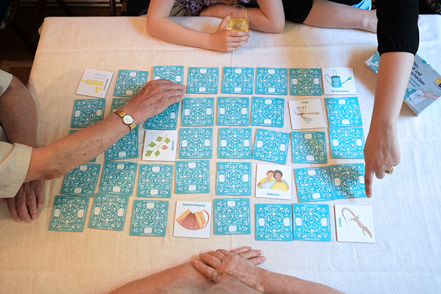Die Karten des Mundart-Memospieles sind auf einem Tisch aufgebreitet. Die meisten Karten sind verdeckt. Nur ein paar Karten liegen offen auf dme Tisch. Rund um den Tisch sitzt eine Familie beim Spiele-Nachmittag.