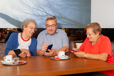 Auf dem Bild sieht man drei ältere Personen, die sich in Kaffeehaus-Atmosphäre über ihre Smartphones austauschen.