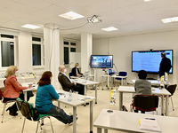 Seminarsituation - Teilnehmer und Teilnehmerinnen schauen nach vorne zum Moderator. Dieser steht vor einem großen Bildschirm und berichtet über das Thema „Moderations- und Interaktionsmethoden" 