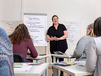 Seminarsituation - Die Referentin steht vor einem Flipchart. Die Teilnehmerinnen sitzen in Reihen vor der Referentin und lauschen aufmerksam.