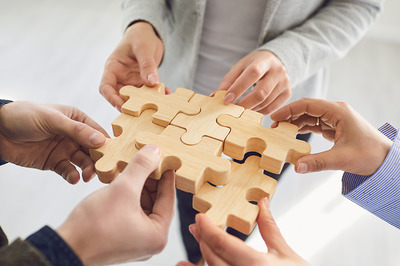 Das Bild die Hände mehrerer Personen, die gemeinsam Puzzle-Teile aus Holz zusammenfügen.