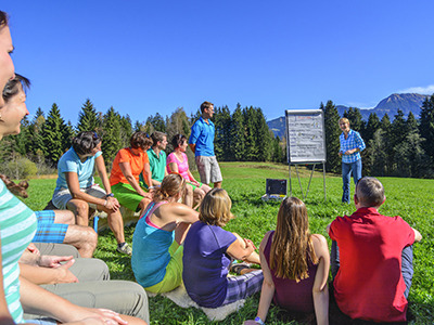 Menschengruppe sitz bei Schönwetter auf einer Wiese und folgen einem Vortrag. Die Vortragende steht vor der Gruppe neben einem Flipchart.. Im Hintergrund sieht man Bäume.