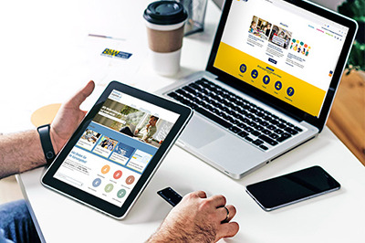 Männerhände halten ein Tablet auf dem die Startseite von https://www.bildungsangebote.at zu sehen ist. Dahinter steht ein Laptop auf einem Schreibtisch, auf diesem sieht man die Startseite der Homepage https://www.bildungsgemeinden-noe.at