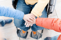 Drei Personen legen die Hände aufeinander. Die Personen tragen bunte Anoraks, eine sitzt im Rollstuhl.