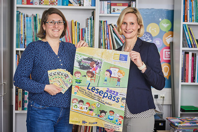 Zeit Punkt Lesen-Projektleiterin Nicole Malina-Urbanz und BhW Niederösterreich-Geschäftsführerin Therese Reinel stehen vor einem Bücherregal und halten ein Plakat der Lesepass-Aktion in die Höhe.