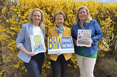 Drei Damen stehen vor einem gelb blühenden Strauch. Zwei der Damen halten je ein Buch in der Hand und die Dame in der Mitte ein Schild mit dem BhW-Logo.