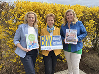 Drei Damen stehen vor einem gelb blühenden Strauch. Zwei der Damen halten je ein Buch in der Hand und die Dame in der Mitte ein Schild mit dem BhW-Logo.