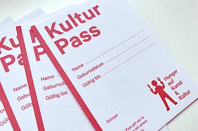 Foto von mehreren Kulturpässen. Es steht auf einem ca. A7 großen Karton, in roter Schrift, Kulturpass, dann Felder zum Ausfüllen für Name und Geburtsdatum. Und man sieht das Logo: das besteht aus einem roten Männchen mit Messer und Gabel in der Hand und daneben steht: Hunger auf Kunst & Kultur