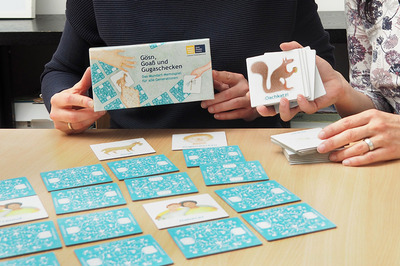Die linken Hände halten die Schachtel des Spiels Mundart-Memo „Gösn, Goaß & Gugaschecken“ , die rechten Hände halten ein paar Memokarten. Auf dem Tisch, sind die Spielkarten aufgelegt.