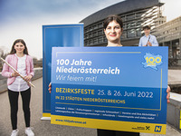 Eine Frau steht im Vordergrund mit einem Schild in der Hand, auf dem für die Bezirksfeste am 25. und 26. Juni geworben wird. Links davon steht eine junge Frau mit Querflöte und weiter im Hintergrund steht ein junger Mann mit Klarinettte. Das Foto wurde vor dem Landhaus St. Pölten aufgenommen.