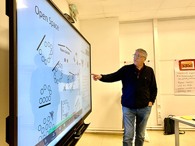 Man sieht den große Bildschirm mit Infos zum Thema "Open Space". Referent Leo Baumfeld zeigt mit deiner rechten Hand auf die Imformationen.
