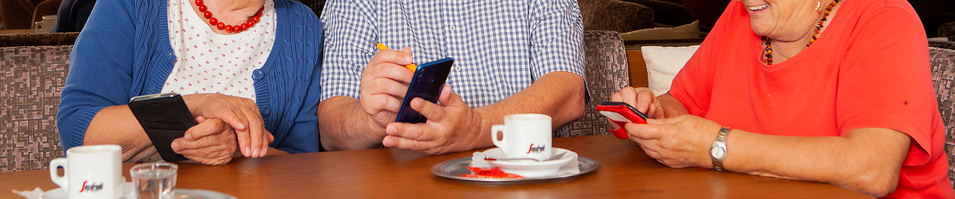 Auf dem Bild sieht man drei ältere Personen, die sich in Kaffeehaus-Atmosphäre über ihre Smartphones austauschen.