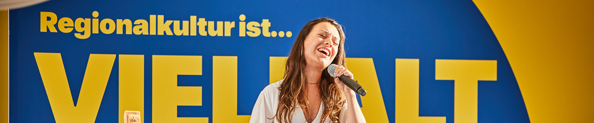 Eine Frau im weißen Kleid steht auf der Bühne und singt in ein Mikrofon.