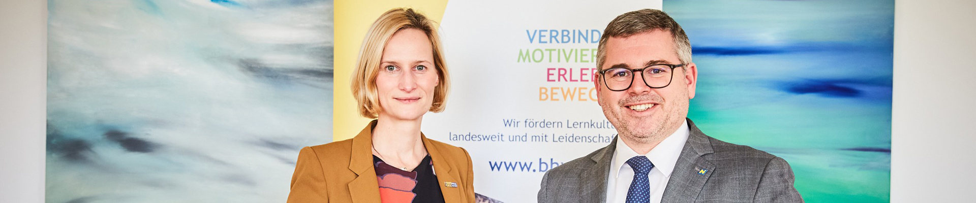 Landesrat Ludwig Schleritzko und BhW Geschäftsführerin Therese Reinel stehen vor einem BhW Rollup und halten die Broschüre "Bildungsagenda NÖ" in den Händen.